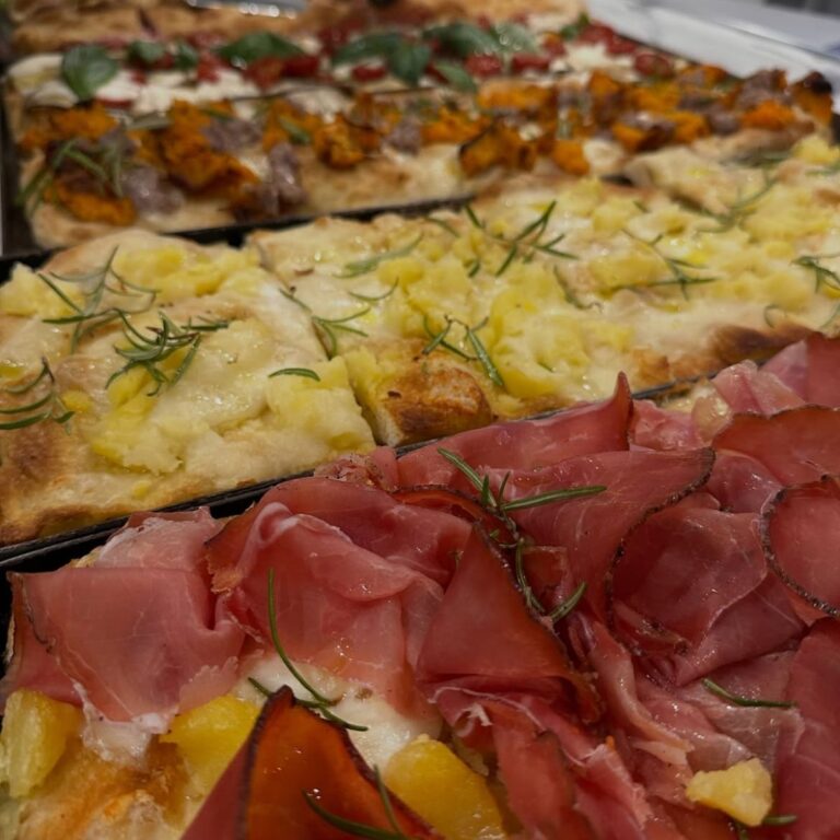 Tranci di pizza gourmet in teglia farciti con salumi, patate, rosmarino e pomodorini in esposizione.