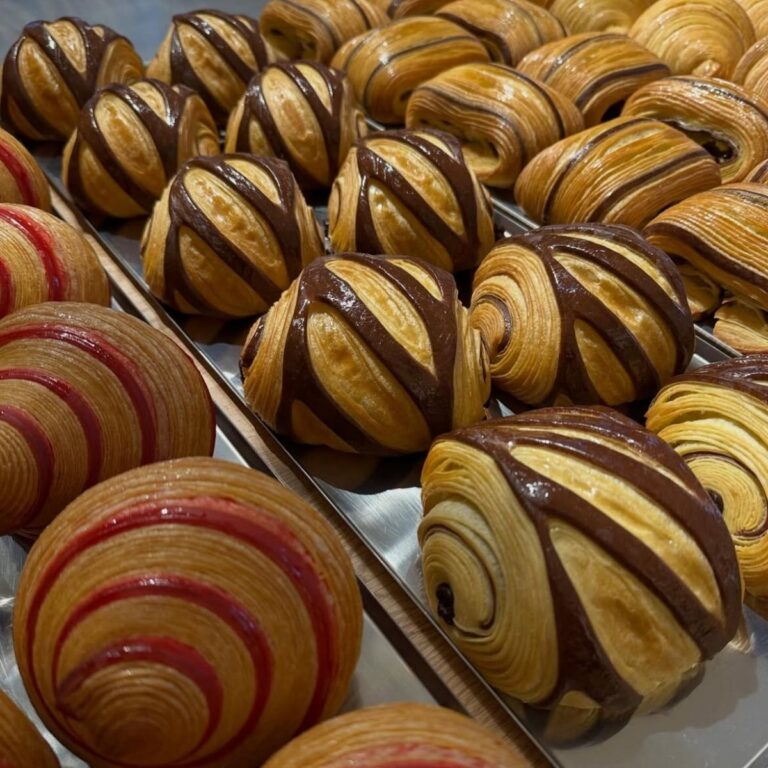 Vassoio di dolci sfogliati da colazione tra cui pain au chocolat bicolore e croissant bicolore ai lamponi perfettamente alveolati.