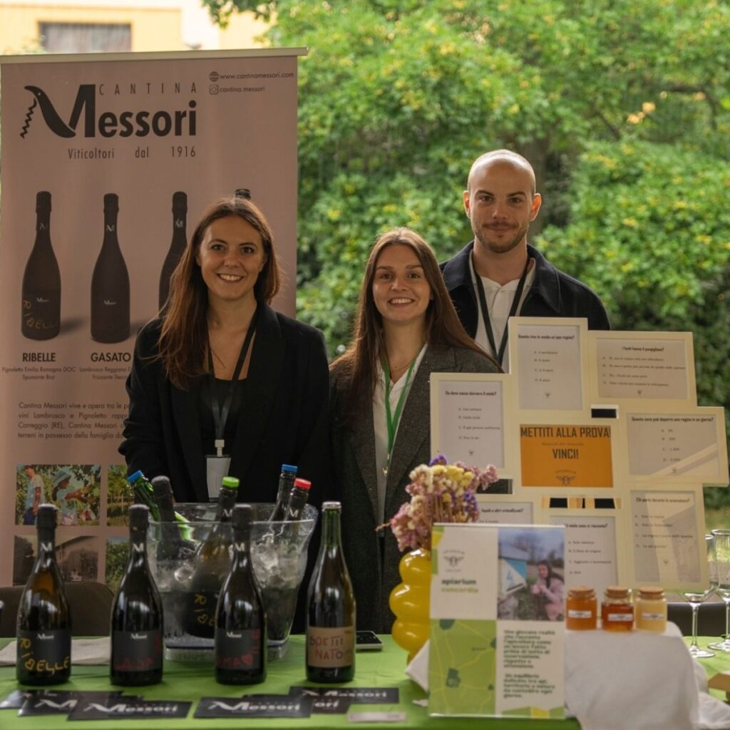 Tre giovani rappresentanti di Cantina Messori sorridenti dietro allo stand di degustazione con bottiglie di vino in primo piano.