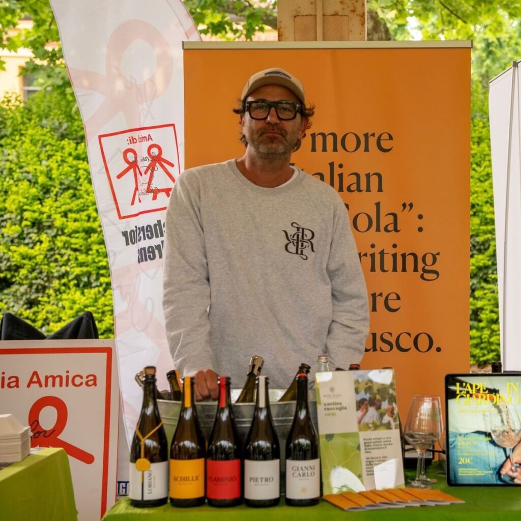 Vignaiolo di Cantina Roncaglia in occhiali e cappellino dietro al bancone con le bottiglie di vino artigianale in primo piano.
