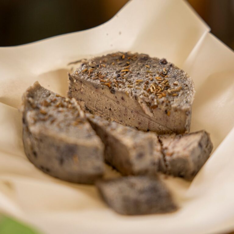 Formaggetta vegetale di anacardi fermentati tagliata a fette su carta alimentare con erbe aromatiche in superficie.