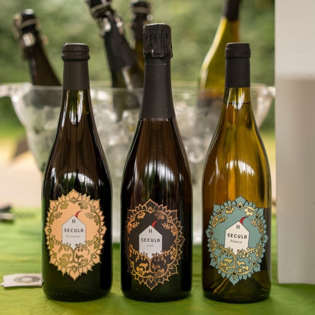 Tre bottiglie di vino Secula con etichette decorative a mandala (Frizzante, Rosé e Bianco) disposte su un tavolo di Freschissimo.