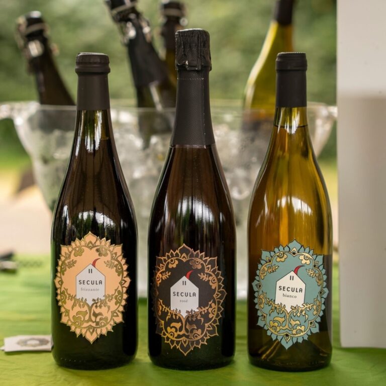 Tre bottiglie di vino Secula con etichette decorative a mandala (Frizzante, Rosé e Bianco) disposte su un tavolo di Freschissimo.