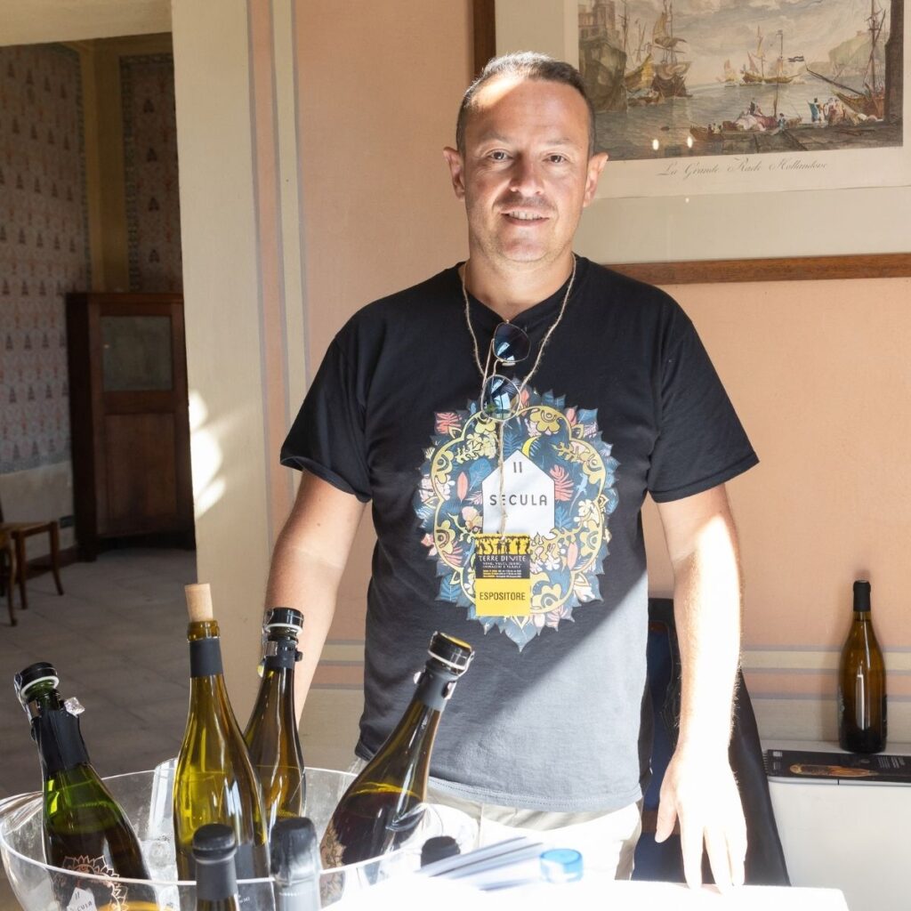 Produttore di Secula in t-shirt personalizzata sorridente dietro al banco di degustazione con bottiglie in primo piano.