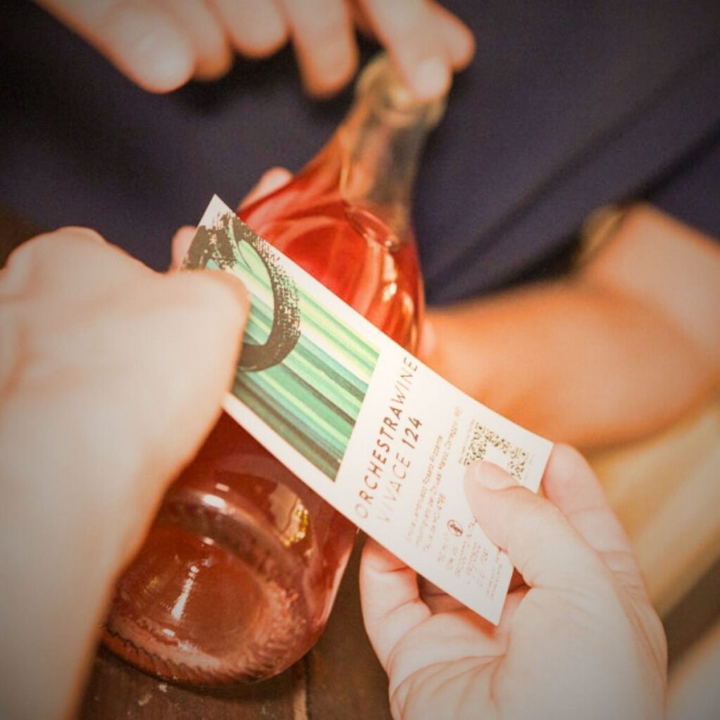 Mani che applicano con cura l'etichetta del vino rosato Vivace 124 di Orchestra Wine sulla bottiglia.