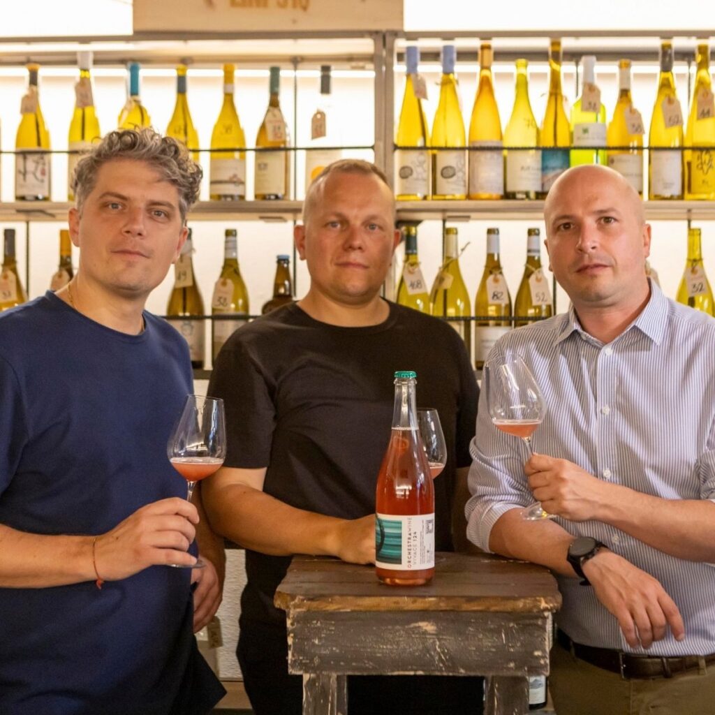 Tre membri del team di Orchestra Wine sorridenti che fanno il gesto del pollice in su dietro al banco di degustazione del festival Freschissimo.