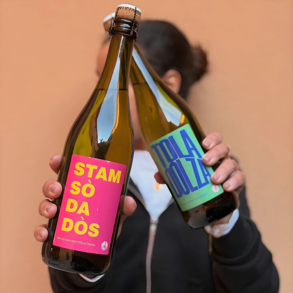 Mani che mostrano due bottiglie di vino di Cantina Bassoli con etichette colorate "Stam Sò Da Dòs" e "Tola Dolza".