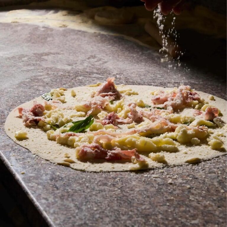 Mani del pizzaiolo che spolverano formaggio grattugiato su un disco di pizza crudo farcito con mozzarella, prosciutti e basilico fresco.