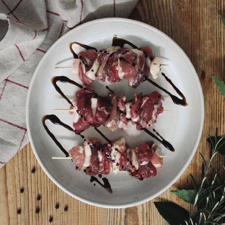 Tre spiedini di carne fresca e lardellata disposti su un piatto bianco e decorati con riduzione di aceto balsamico.